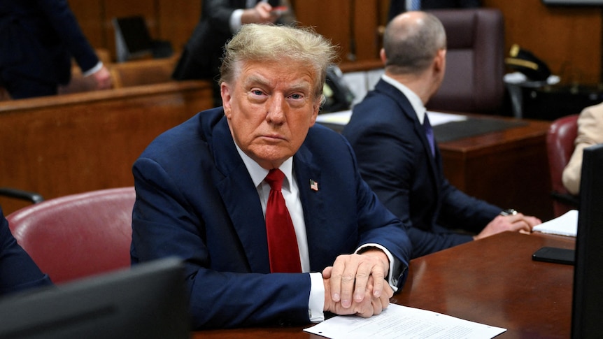 Donald Trump site in a courthouse, looking directly at the camera with a solemn expression.