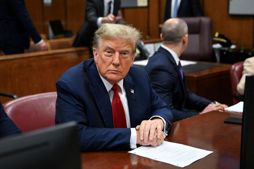 Donald Trump site in a courthouse, looking directly at the camera with a solemn expression.