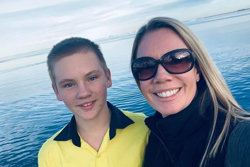 Trinity Rafferty, wearing sunglasses, alongside her son with the ocean in the background.