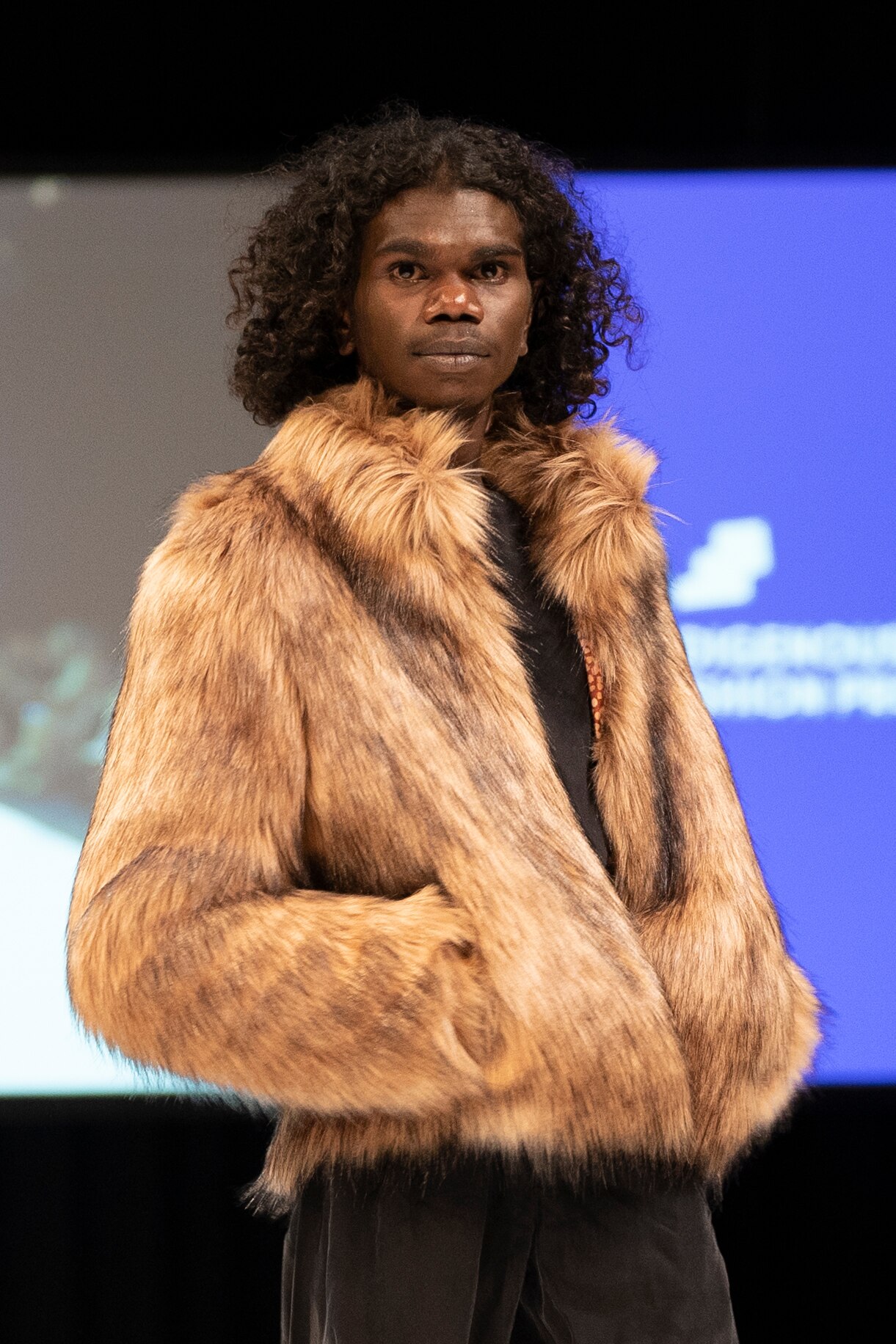 A man with curly hair looks into the camera.  He is wearing a fur jacket. 