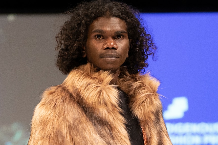 A man with curly hair looks into the camera. He is wearing a fur jacket. 