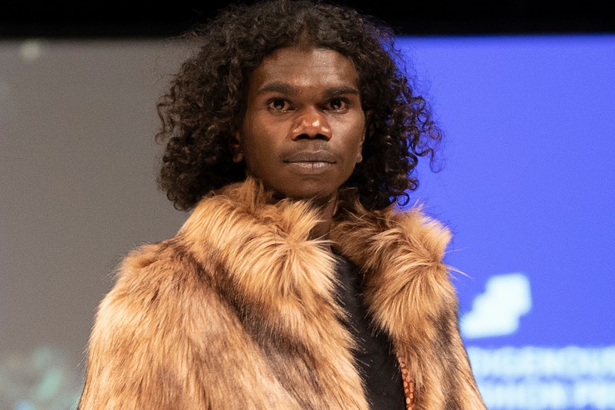 A man with curly hair looks into the camera.  He is wearing a fur jacket. 