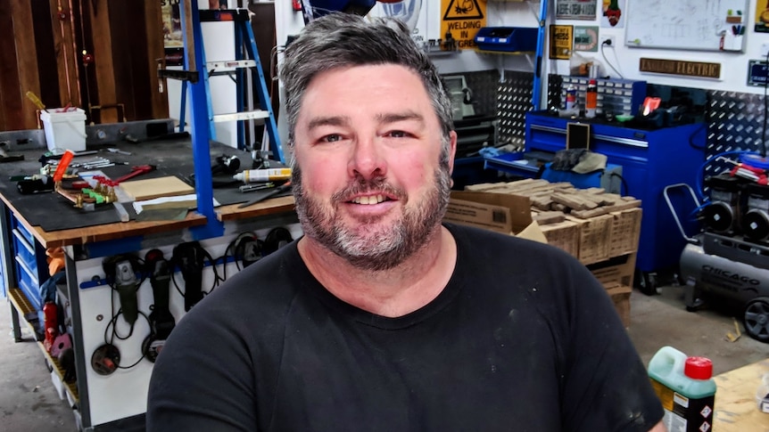 A middle-aged man in a garage full of tools and machinery. 