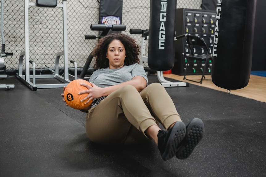 A woman with brown skin is exercising in a gym using a weighted ball.