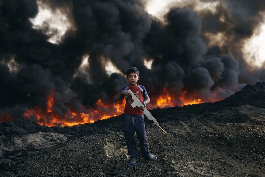 A boy stands in front of burning oil fields.