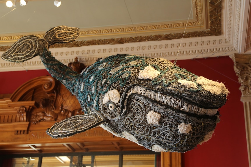Large baleen whale made of fabric, rope and material suspended from room of ornate old room.