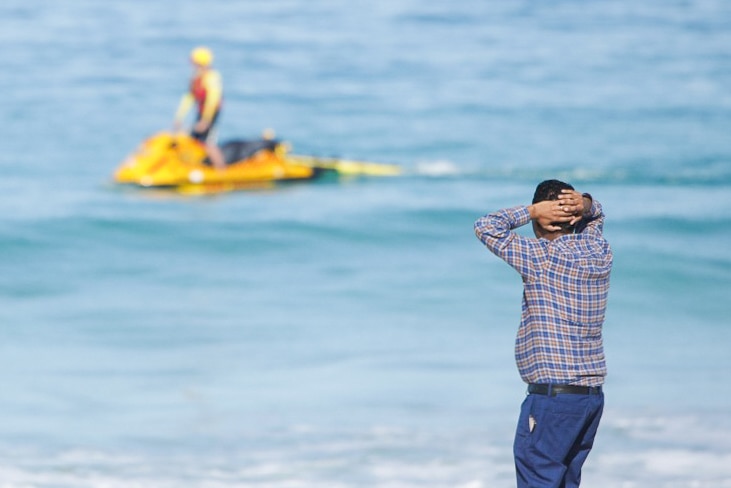 A man with his hands on his head looks out over the ocean with a search jetski on the water in the background.