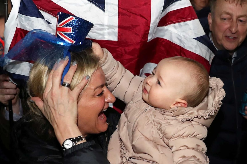 A smiling woman holds a baby in front of the Union Flag.