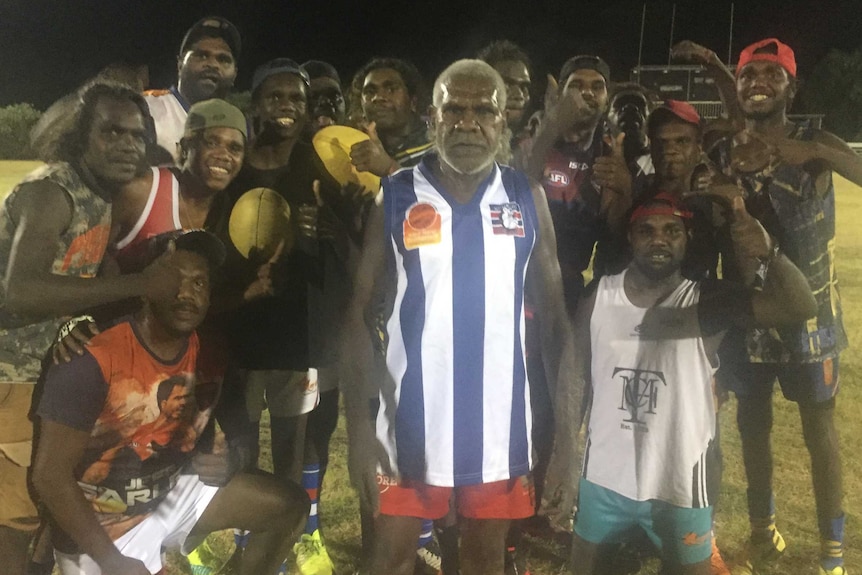 AFL player poses for the camera before other men cheering him on. Nighttime on an oval.