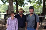 Three people in riding gear smile for the camera in front of a pen of horses.