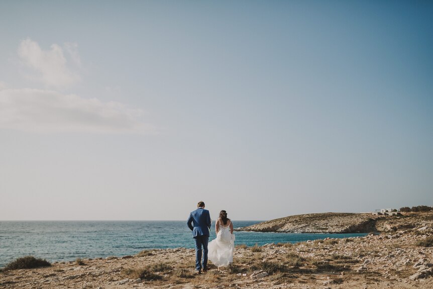 A bride and groom in wedding dress seen from the back walking across rocks with the blue ocean in front of them