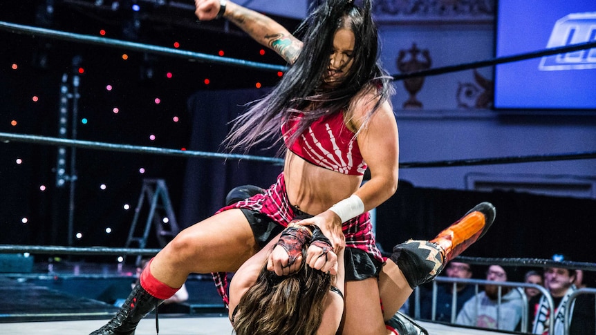 Female wrestler Kellyanne sitting on and raising right hand over wrestling opponent.