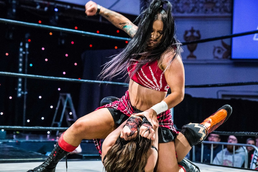 Female wrestler Kellyanne sitting on and raising right hand over wrestling opponent.