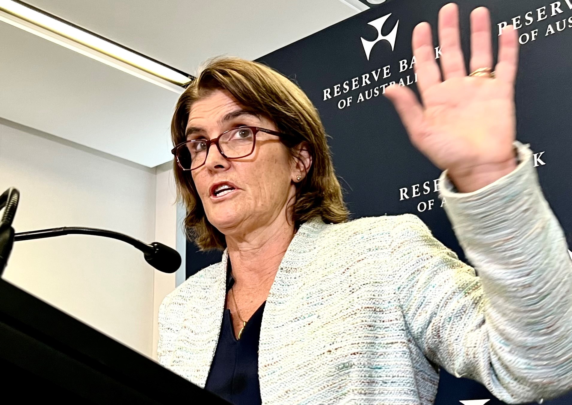 RBA governor Michele Bullock holds her hand up while speaking at a press conference.