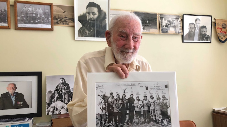 An elderly man holds a black-and-white photo of men in Antarctic, in front of other black and white photos on a wall.