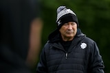 A man in black cold weather gear watches football training.