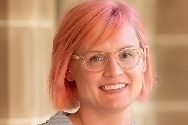 Una mujer con cabello rosado muerto y gafas de pie frente a la cámara.