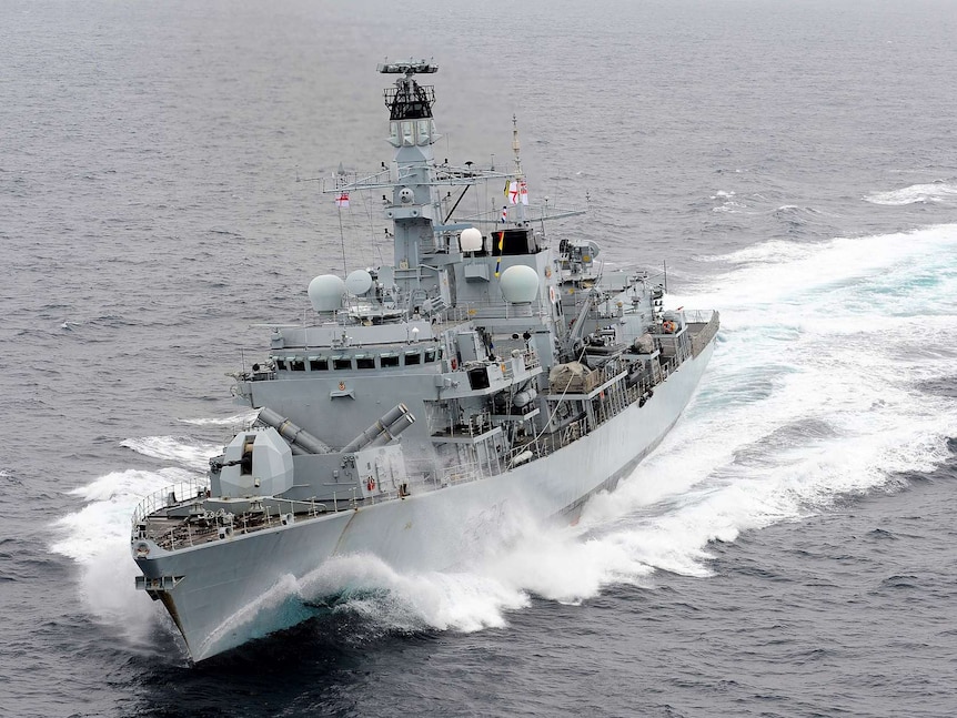 A grey Royal Navy ship cuts through grey sea waters on an overcast day.