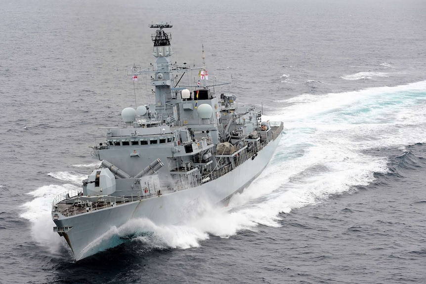 A grey Royal Navy ship cuts through grey sea waters on an overcast day.