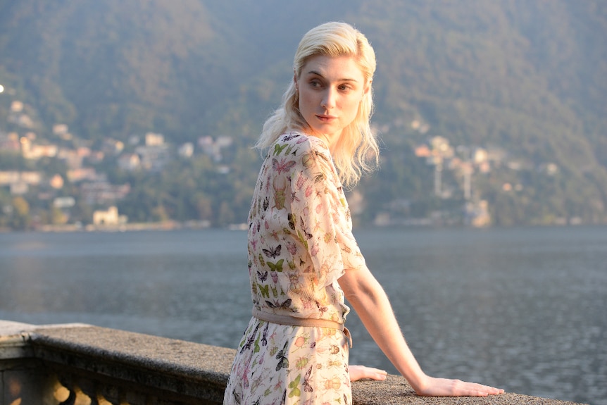 A young blonde woman in long dress decorated with insect illustrations and stands by lake's edge looking back, sun on her face.