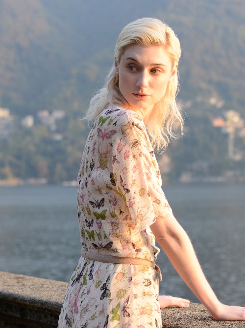 A young blonde woman in long dress decorated with insect illustrations and stands by lake's edge looking back, sun on her face.