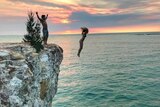 Young people jumping from rocks into open water at sunset.