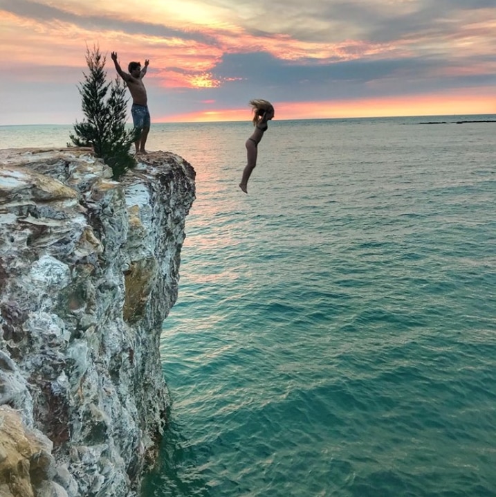 Young people jumping from rocks into open water at sunset.