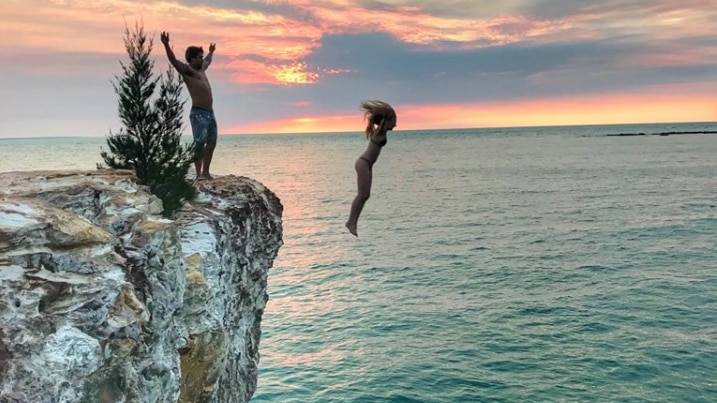 Young people jumping from rocks into open water at sunset.