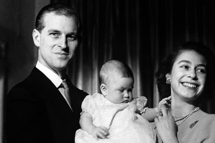 A black and white photo of Prince Philip in a suit holding baby Prince Charles while the Queen looks off to the side.