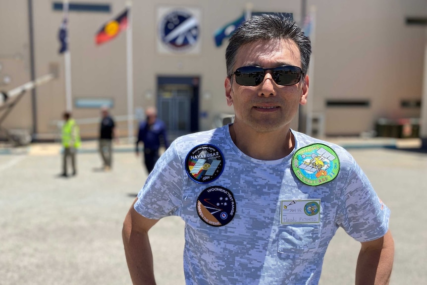 A nab wearing sunglasses and a blue T-shirt with badges on it in front of a large building