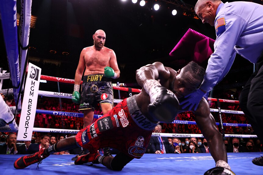Deyontay Wilder falls face first to the ground with a man in a blue shirt trying to catch him and Tyson Fury watching on