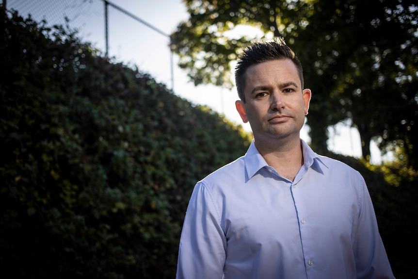 A man in a blue shirt stands in front of a hedge