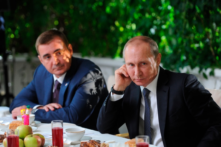 Vladimir Putin rests his hand on his chin while sitting at a table with another man in a suit. There is fruit on the table