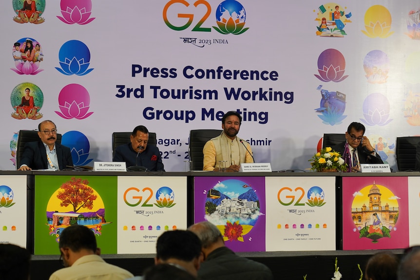 A group of four men sit behind a desk in front of a sign saying, Press Conference 3rd Tourism Working Group Meeting