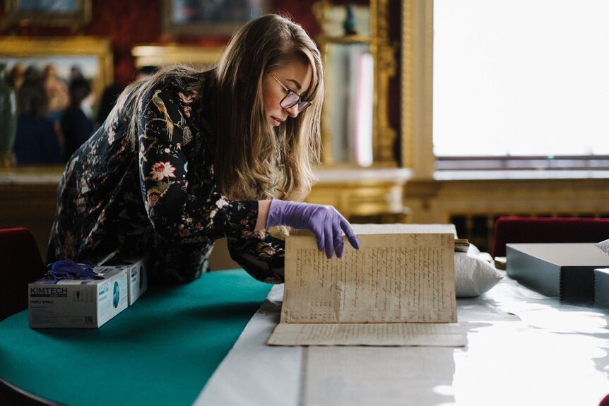 A young woman with glasses, wearing gloves, carefully opens a very old book