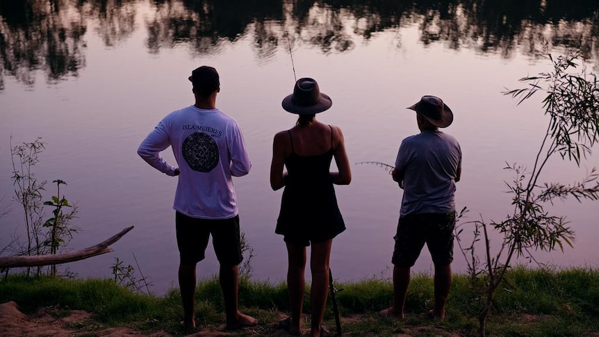 Three people, seen in silhouette, standing and fishing on the banks of a river in a purple dusk.