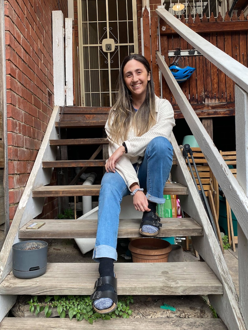 Becc Brooker sits smiling on the front steps of a brick home.