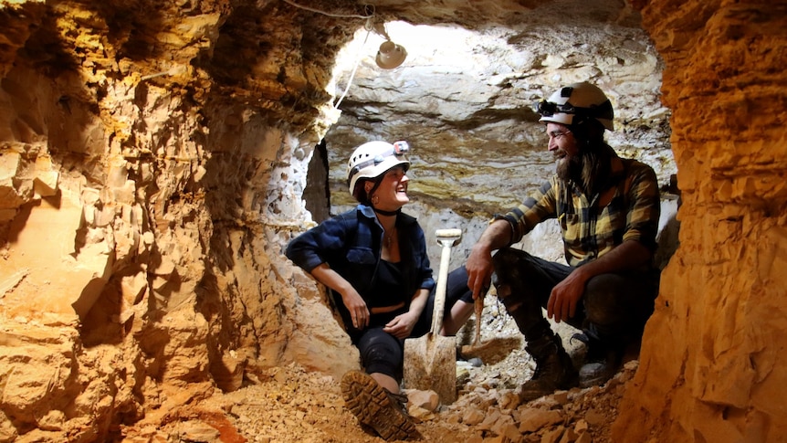 Two people wearing hard hats sit with shovels and tools, looking at each other surrounded by rock walls. 