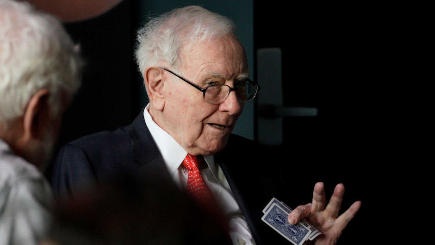 Investor Warren Buffett in a black suit with a red tie, holding playing cards in his hand.