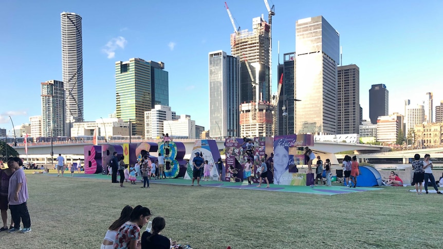 Three young women sitting on a picnic rug on grass in front of a large, multi-coloured sign that spells out "Brisbane"