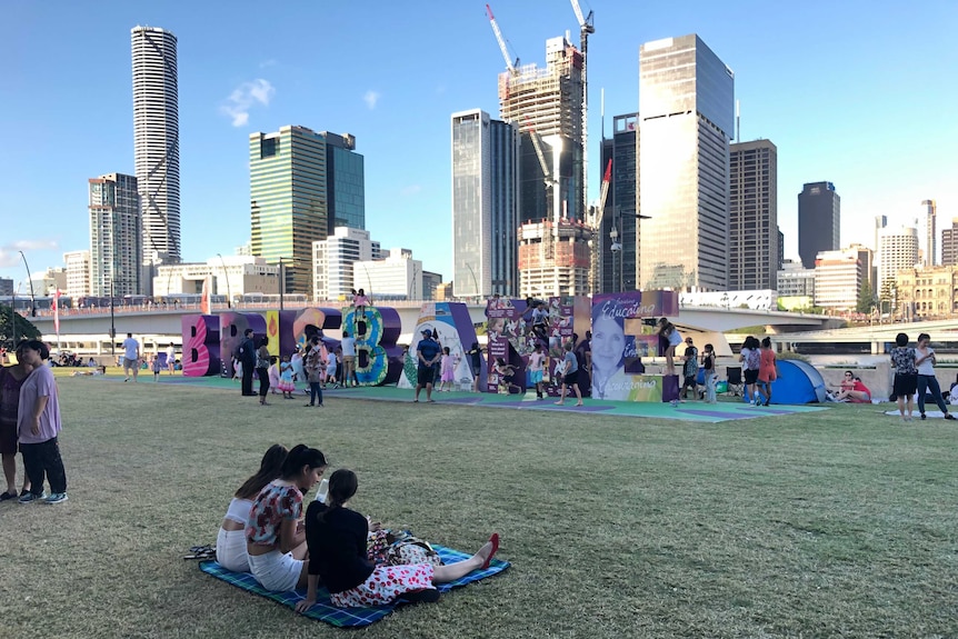 Three young women sitting on a picnic rug on grass in front of a large, multi-coloured sign that spells out "Brisbane"