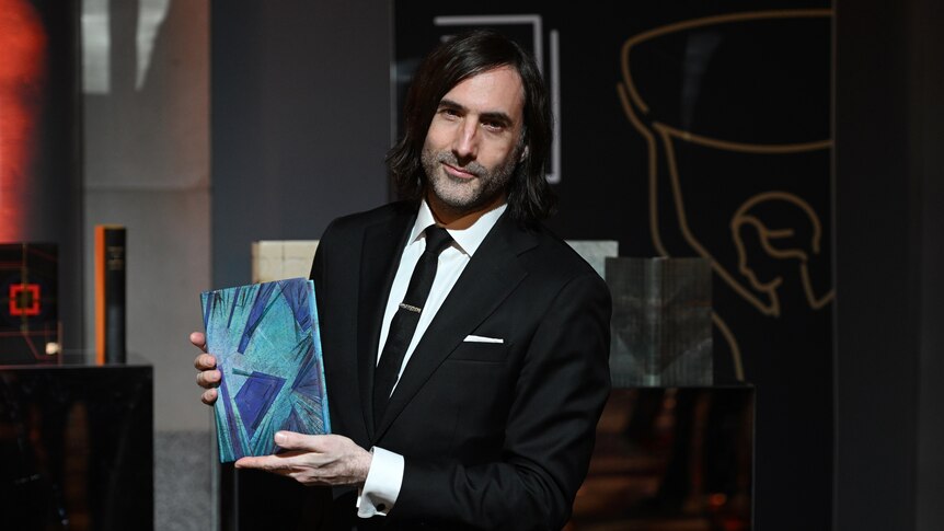 A white man with dark, chin-length hair, wearing a suit, holds a book up to the camera