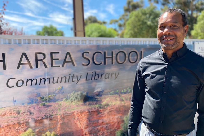 A man with dark skin has a greying beard and short hair, he wears a dark shirt in front of a school sign