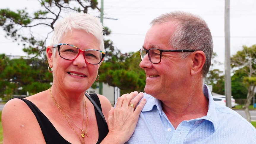 Woman with short white hair and glasses looks at camera as her hand rests on the shoulder of a man with grey hair and glasses