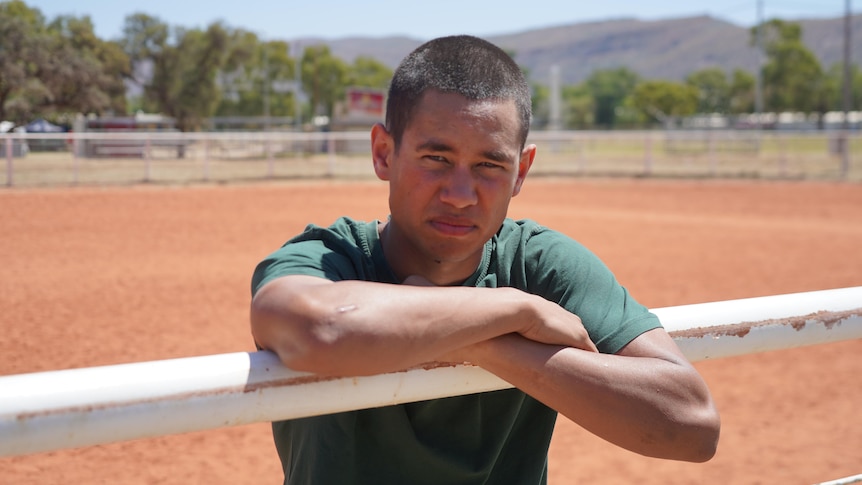 A young Aboriginal man leans forward on the white rodeo arena railing and stares at the camera. He wears green prison garb.