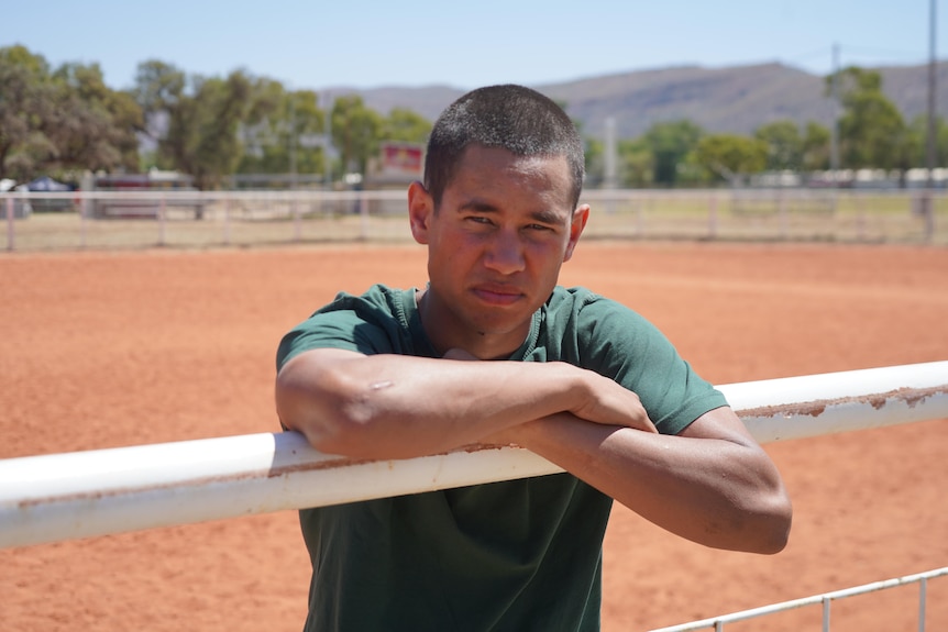 A young Aboriginal man leans forward on the white rodeo arena railing and stares at the camera. He wears green prison garb.