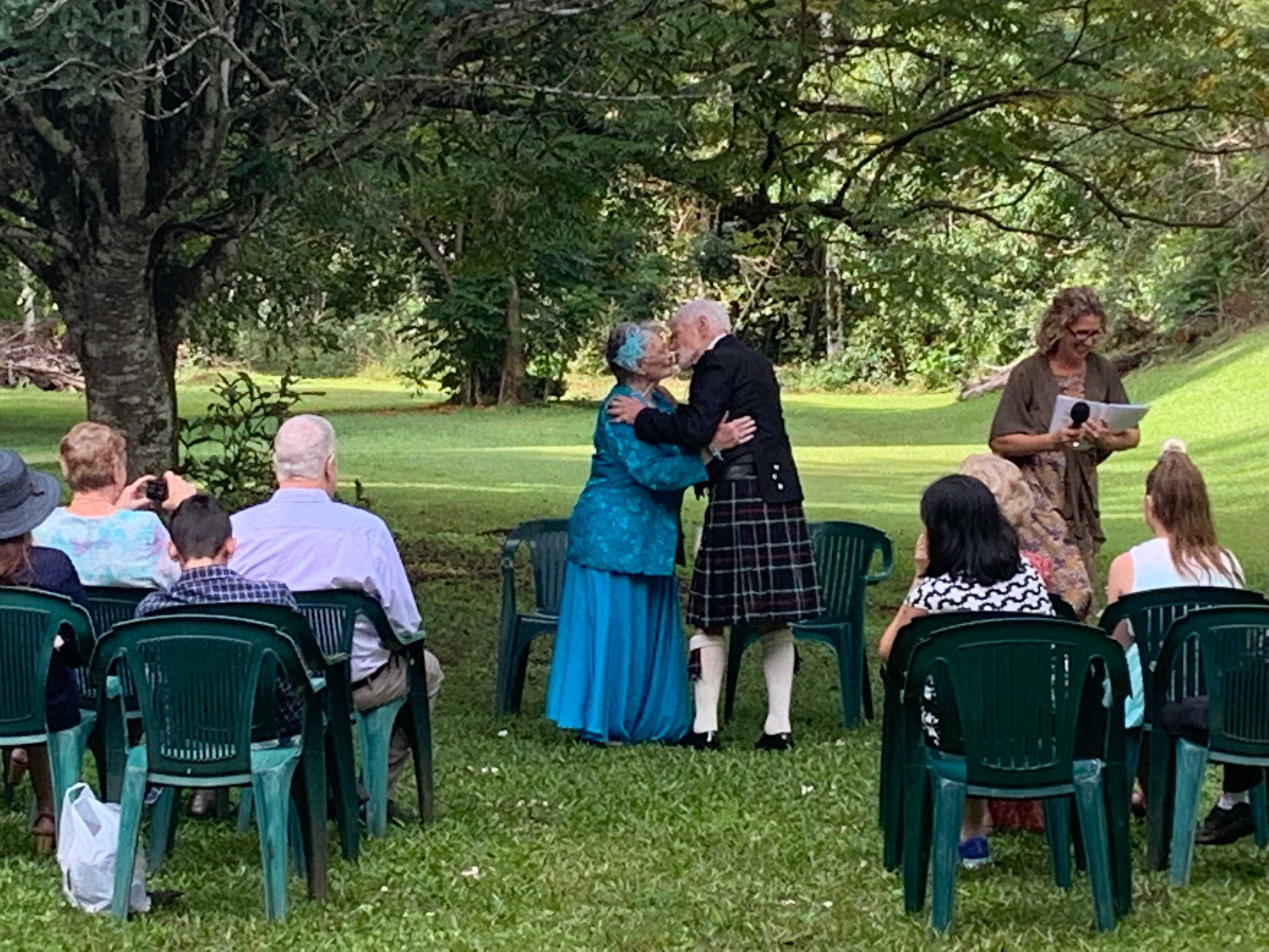 elderly woman in blue dress kissing elderly man in Scottish kilt in a park setting with people in chair watching