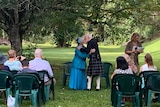elderly woman in blue dress kissing elderly man in Scottish kilt in a park setting with people in chair watching