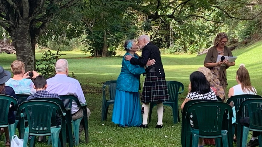 elderly woman in blue dress kissing elderly man in Scottish kilt in a park setting with people in chair watching