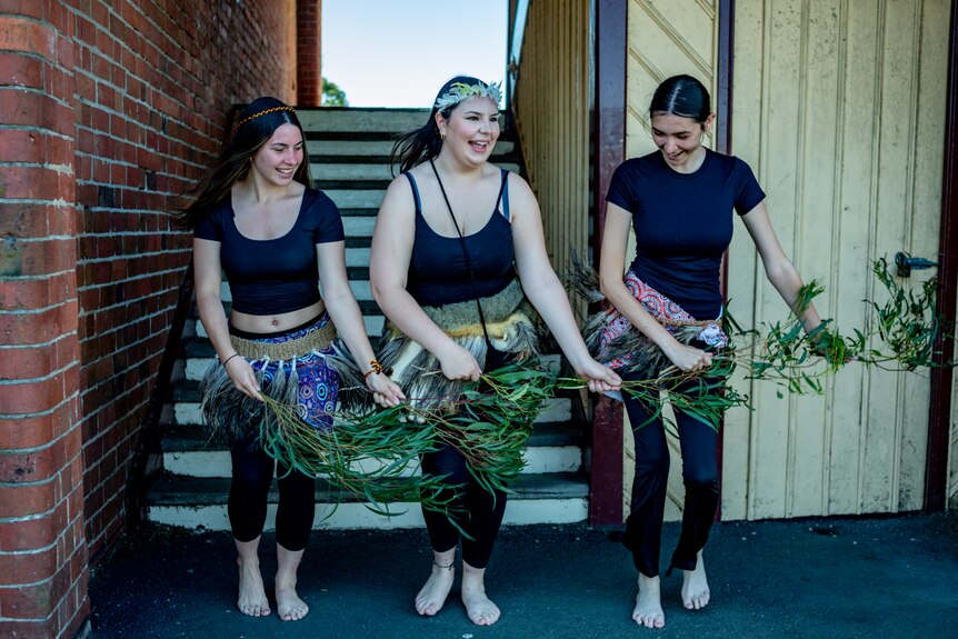 Three girls wearing traditional Indigenous feathered skirts hold branches as they dance.